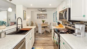 A compact kitchen with white cabinets, a central island, a dining area with wicker chairs, and a ceiling fan.