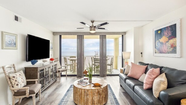 A modern living room with a flat-screen TV, a wooden coffee table, a leather sofa, and a balcony with a view of the ocean.
