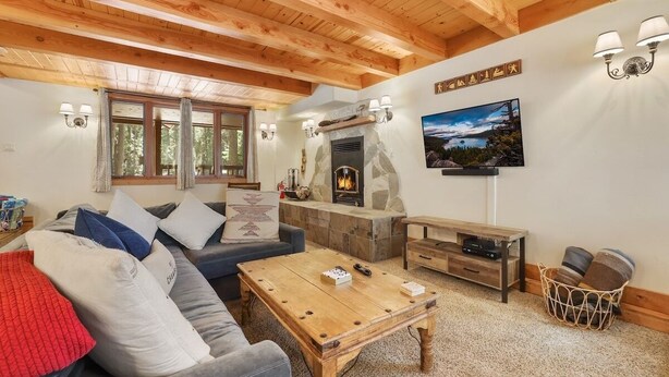 A cozy living room with a stone fireplace, wooden ceiling beams, a sofa with cushions, a coffee table, and a television mounted on the wall.