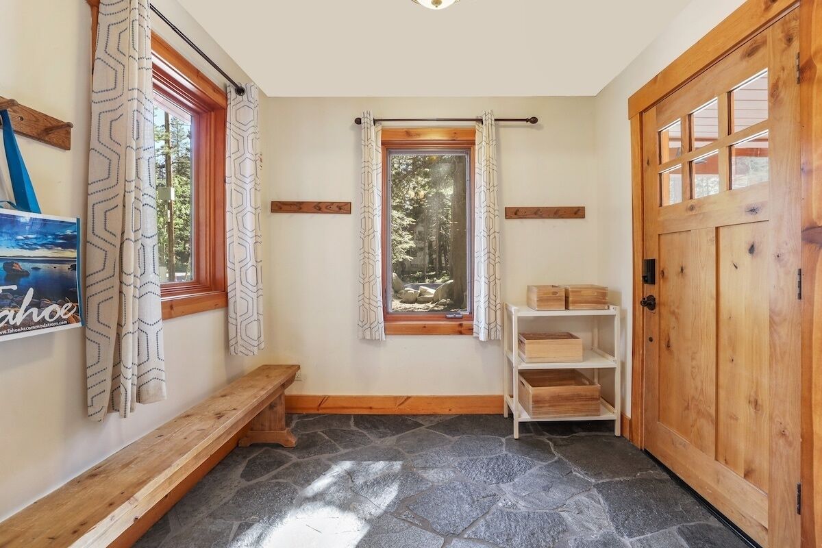 Un pasillo de piedra con suelo, un banco de madera, una puerta de madera y una ventana con cortinas estampadas.