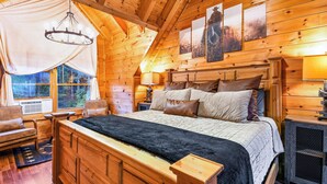 Un dormitorio de cabaña de madera con una cama grande, cabecera de madera, una ventana con cortinas, una mesita de noche y una lámpara.