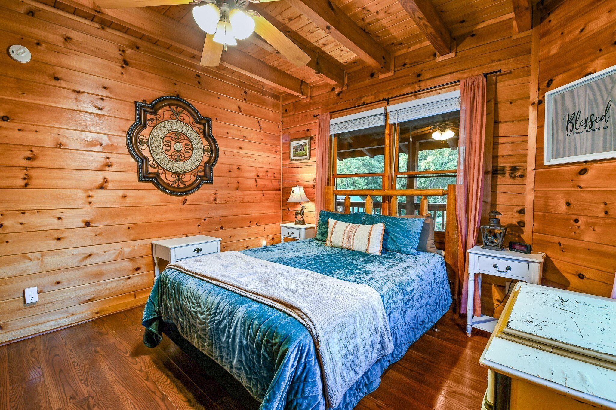 A wooden cabin bedroom with a bed, nightstands, a dresser, and a window with curtains.