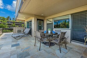 Une terrasse couverte avec un sol en pierre, une table ronde entourée de chaises et une vue sur un paysage tropical, visible à travers de grandes fenêtres.