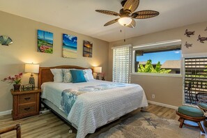 Une chambre à coucher comprenant un lit, des tables de chevet, un ventilateur de plafond, des œuvres d’art accrochées aux murs et une fenêtre donnant sur un paysage agréable.