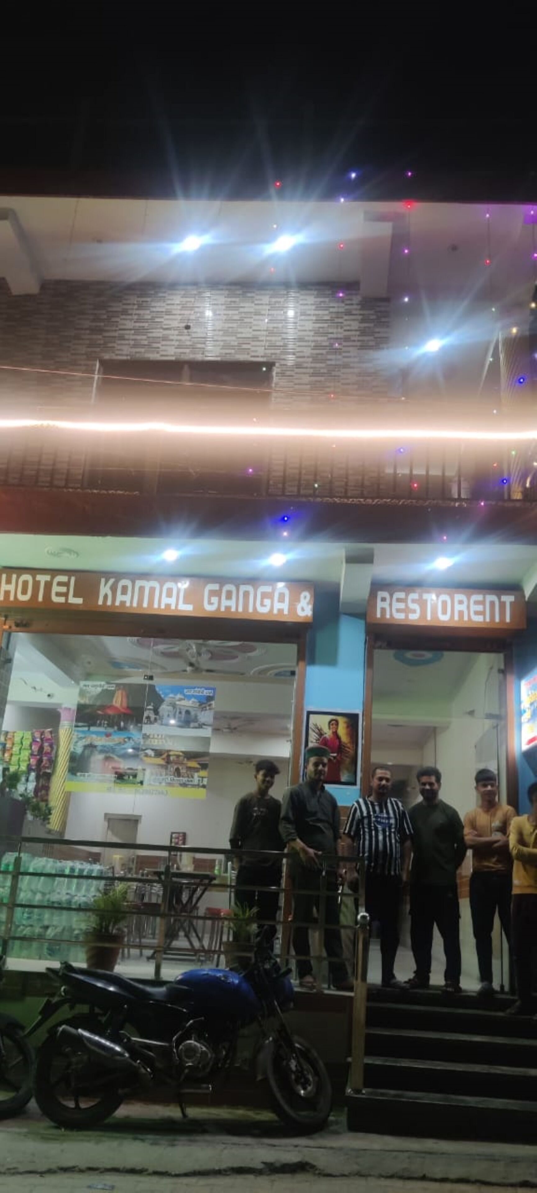 A building with signs for 'HOTEL KAMAL GANGA' and 'RESTORANT'. There are people standing outside, and a motorcycle parked in front.