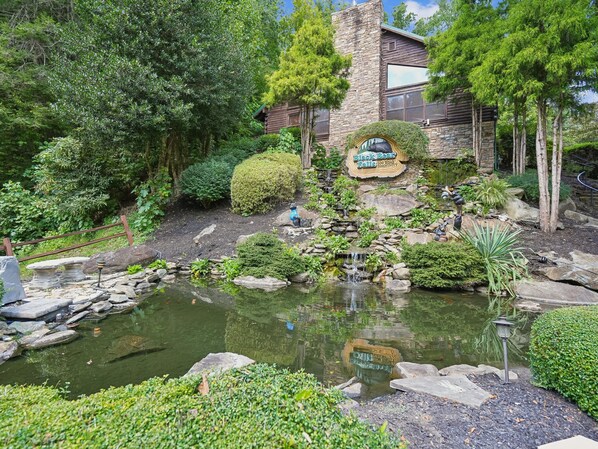 Un jardín ajardinado con un estanque, muros de piedra y una cabaña de madera con la inscripción 'Black Bear Falls'.