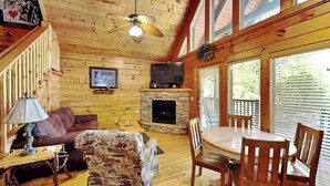 A cozy cabin interior with a wooden floor, a fireplace, a sofa, a dining table with chairs, and a ceiling fan.