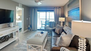 A modern living room with a flat-screen TV, a ceiling fan, a sofa, a coffee table, and a view of the balcony. - Seaside 508 | Updated Interior + Gulf Views (Fort Myers Beach)