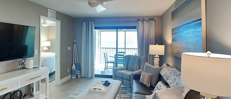 A modern living room with a flat-screen TV, a ceiling fan, a sofa, a coffee table, and a view of the balcony.
