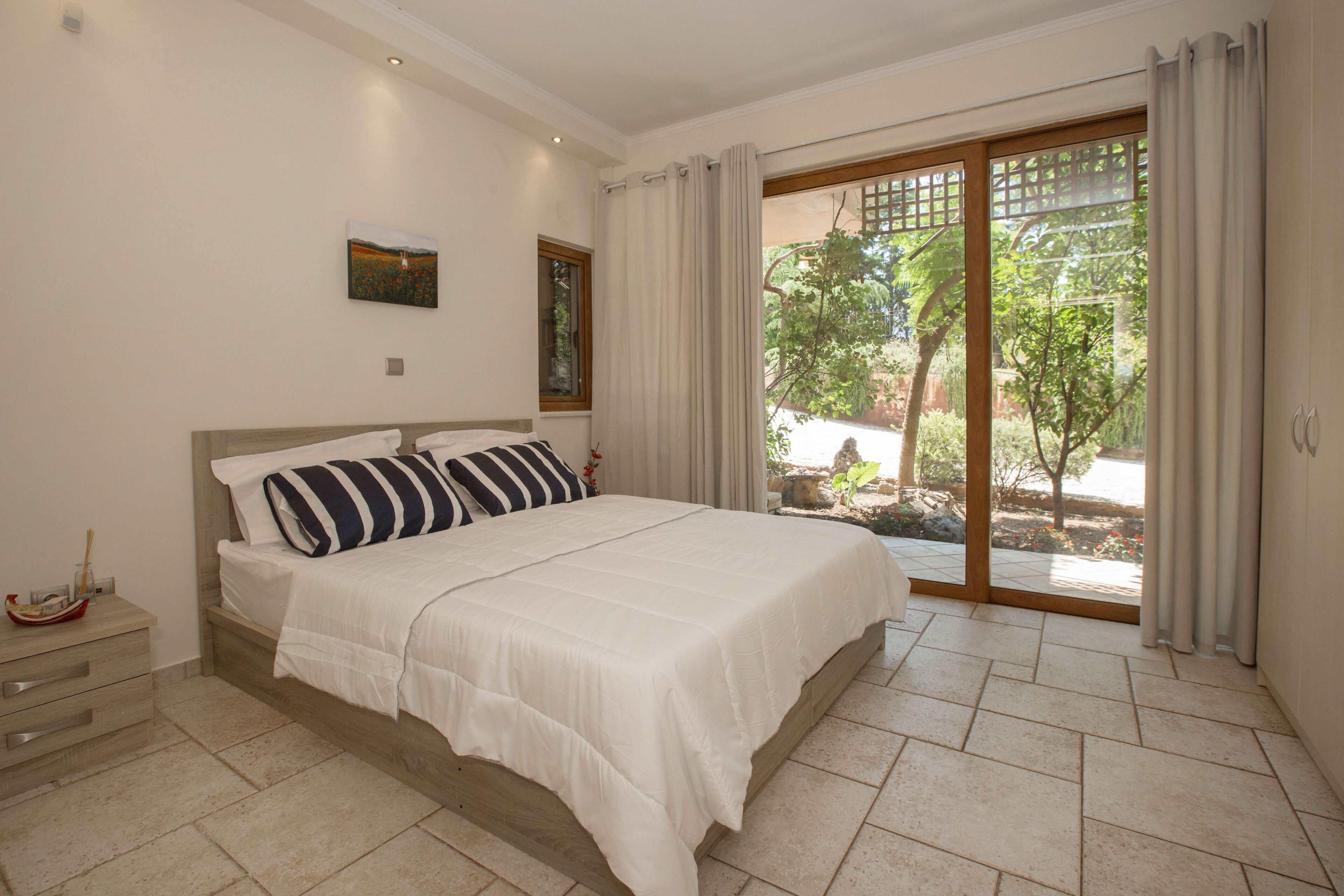 A bedroom with a large bed, a nightstand, a window with curtains, and a view of greenery outside.