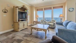A living room with a television, a coffee table, a sofa, and a chair. There is a balcony with a railing and a view of the sky.