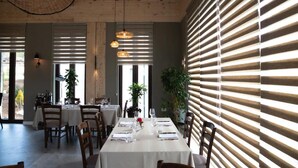 Un coin repas avec une longue table dressée pour quatre personnes, entourée de chaises en bois. Deux lampes suspendues éclairent la table. La pièce est dotée de grandes fenêtres équipées de stores horizontaux, ce qui permet à la lumière naturelle d’entrer.