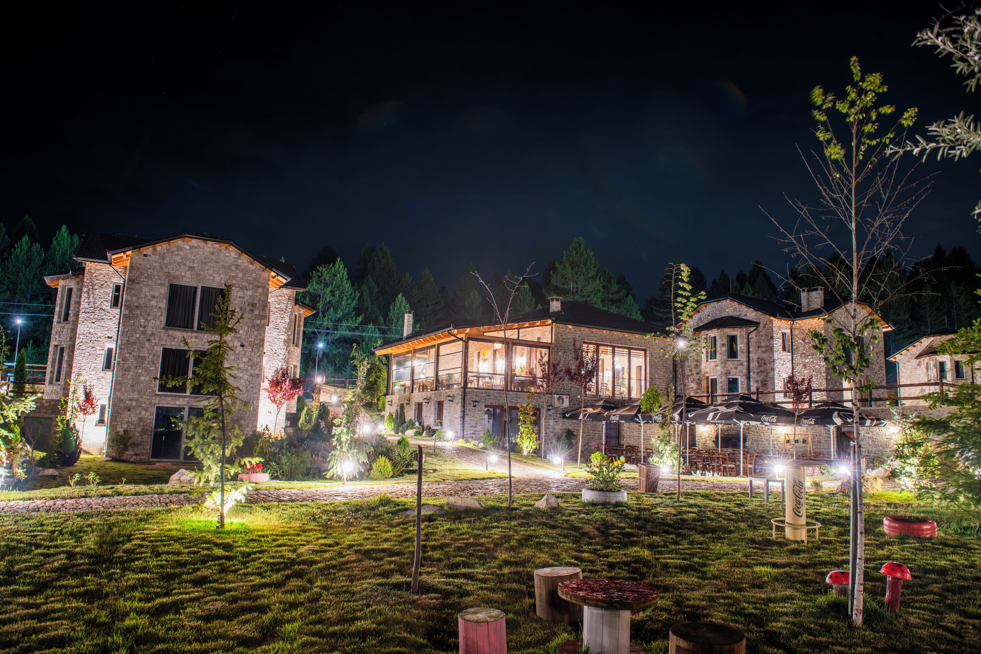 A stone building with a large glass window, surrounded by a well-lit garden with trees and outdoor seating.