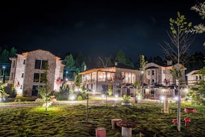Un bâtiment en pierre doté d’une grande fenêtre en verre, entouré d’un jardin bien éclairé, agrémenté d’arbres et de coins salons extérieurs.