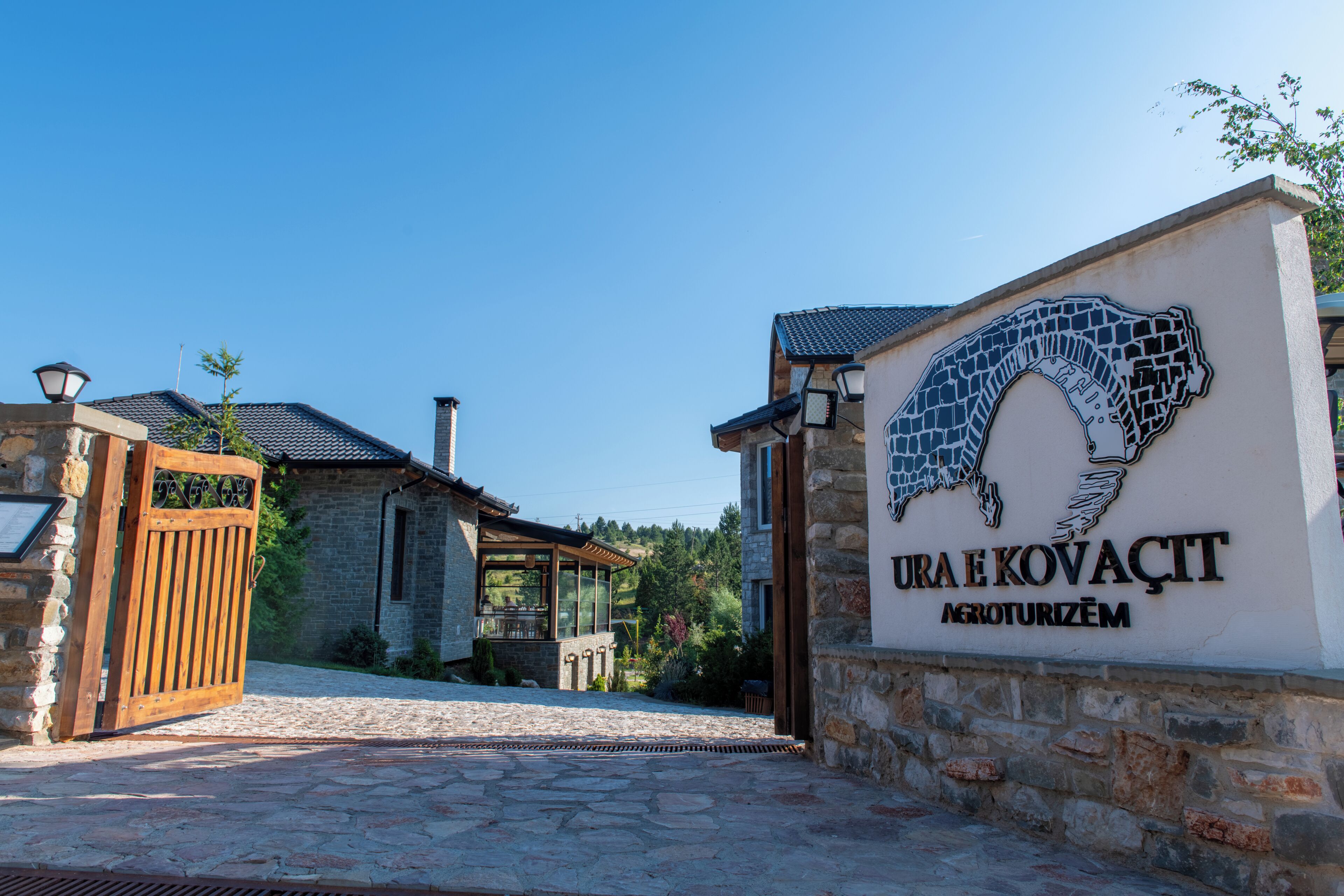 A stone building with a sign that reads 'URA E KOVAĆT' and a wooden gate.
