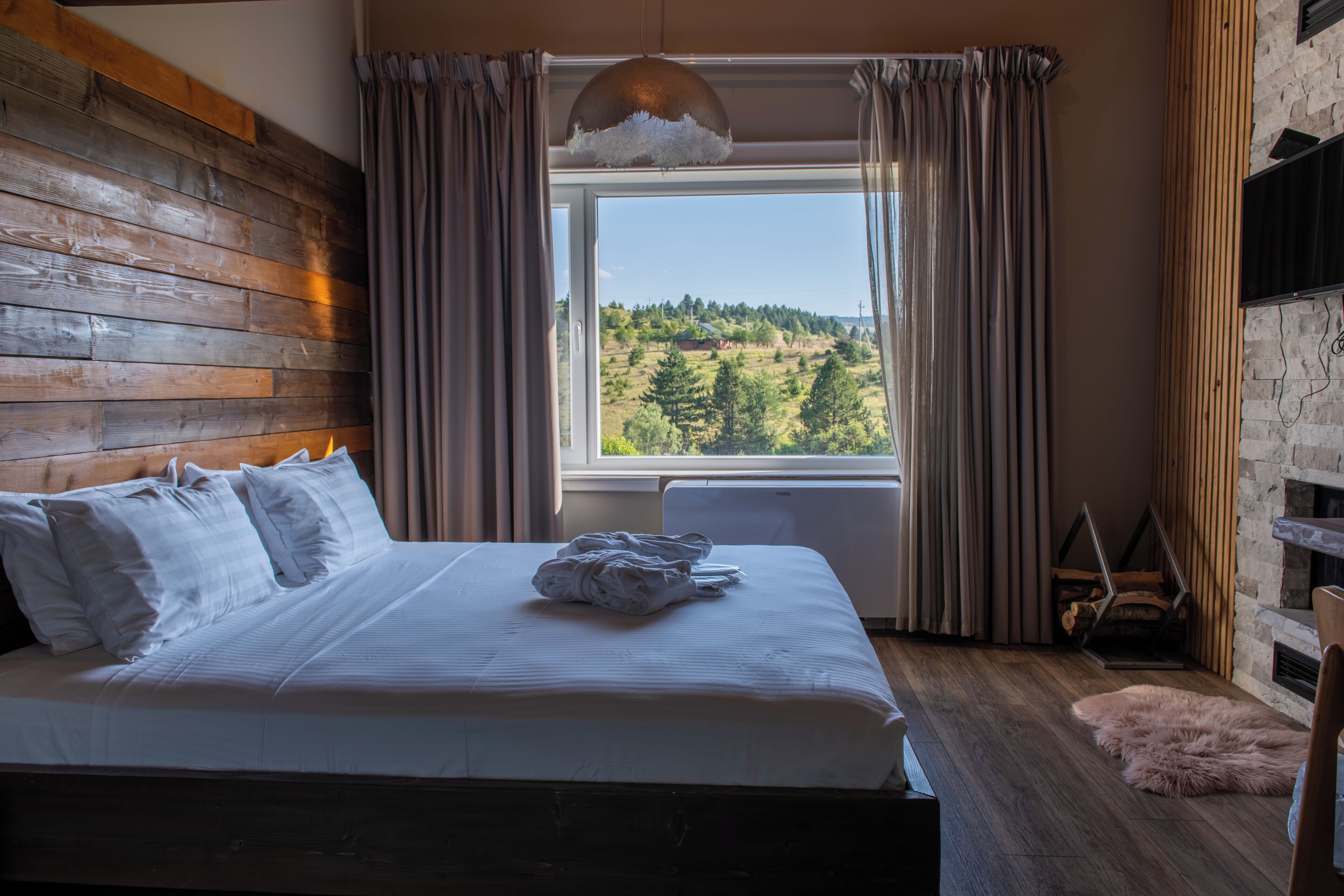 A bedroom with a wooden headboard, a large bed with white bedding, a window with curtains, and a view of greenery outside.