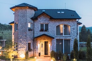 Een twee verdiepingen tellend stenen huis met een torentje, grote ramen en een goed verlichte ingang.