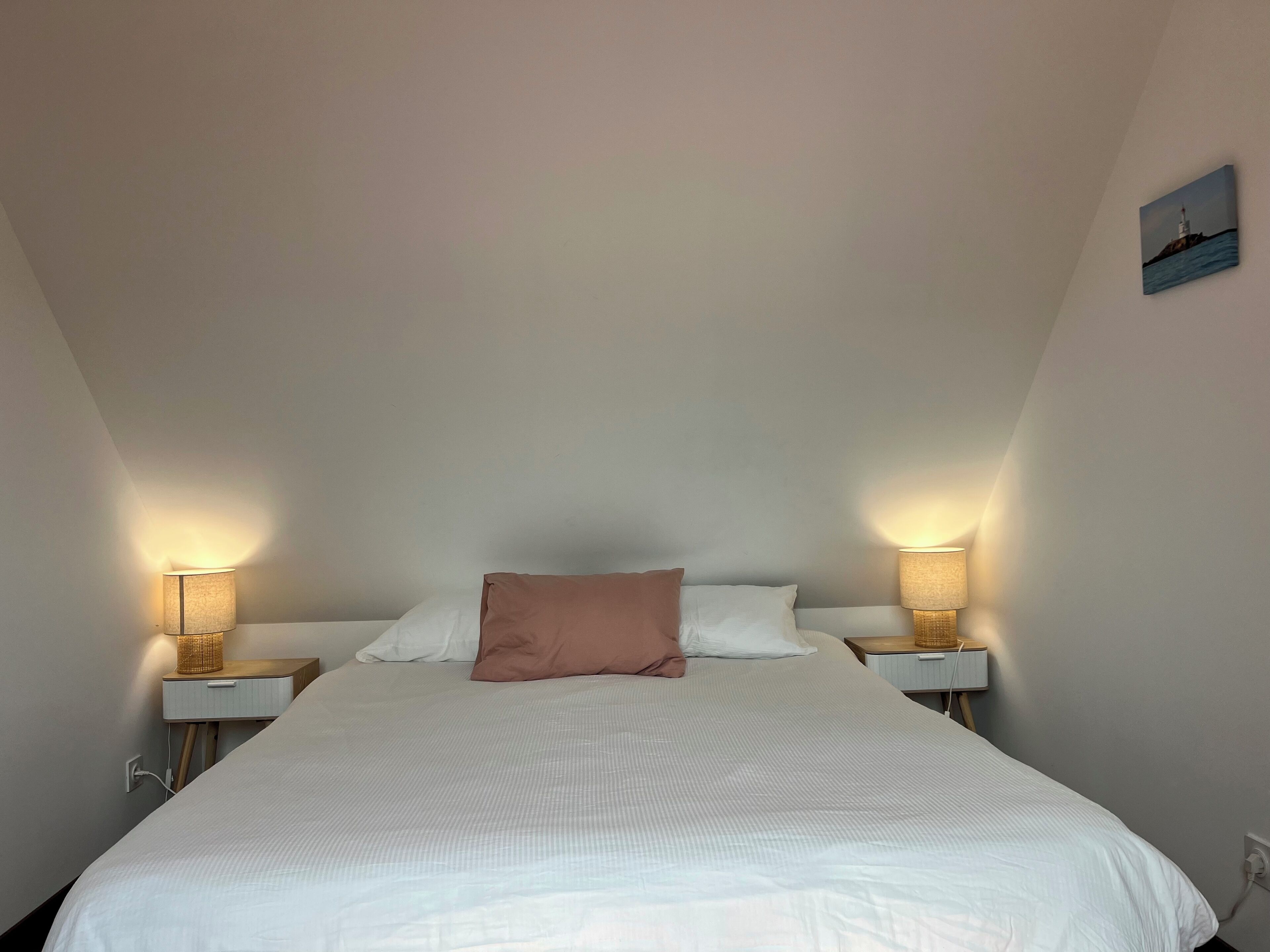 Een net opgemaakt bed met witte lakens en één bruin kussen, met aan beide zijden nachtkastjes met lampen.