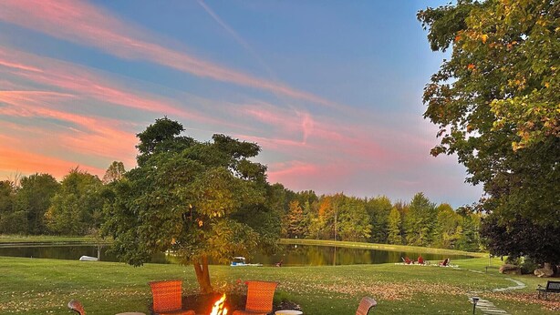 A circular outdoor seating area with a fire pit, surrounded by wicker chairs, set against a backdrop of a lake and trees under a sunset sky.