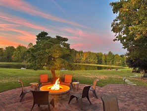Un espace extérieur circulaire aménagé avec des sièges, doté d’un foyer, entouré de fauteuils en osier, le tout se fondant dans un décor composé d’un lac et d’arbres, le ciel se parant des couleurs du coucher de soleil.