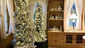 A living room with two decorated Christmas trees, a wooden cabinet with figurines, and a lamp on a side table.