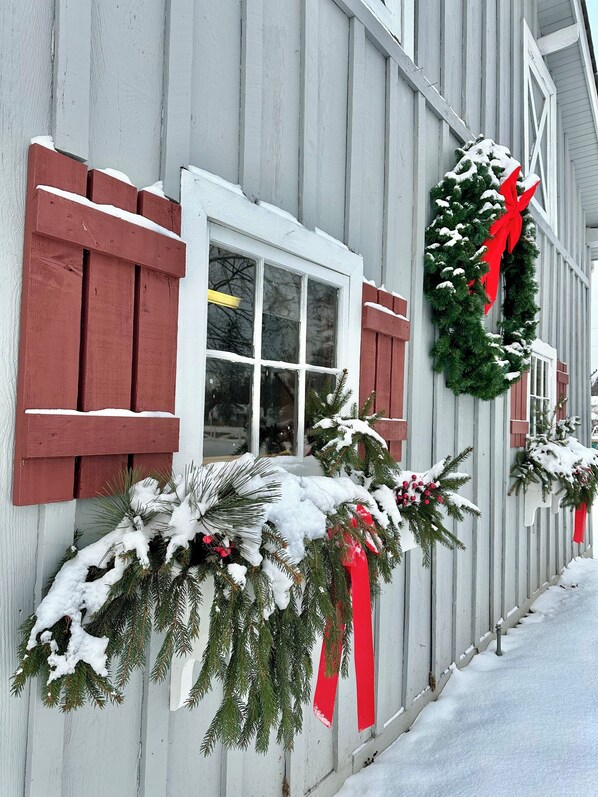 Un edificio con revestimiento blanco y contraventanas rojas, decorado con coronas cubiertas de nieve y ramas de pino.