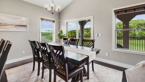 A dining area with a glass-top table, dark wooden chairs, and a chandelier. There are large windows with views of greenery and a pergola. - Fairway Lake Retreat (Camdenton)
