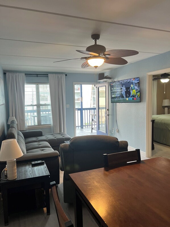A living room with a sofa, a dining table, a TV mounted on the wall, and a ceiling fan.