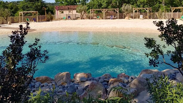 A clear pool surrounded by rocks and trees, with a wooden fence and a small building in the background.