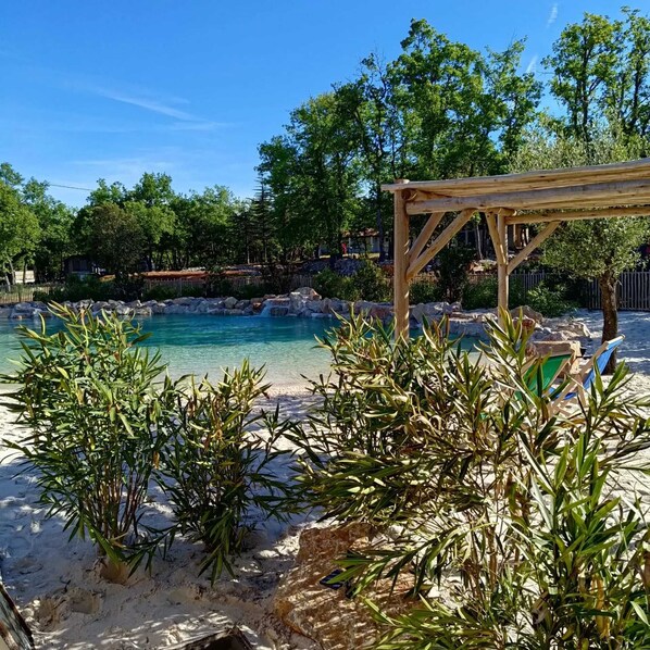 Une piscine aux eaux cristallines, surmontée d’une pergola en bois, et entourée de sable et de plantes tropicales.