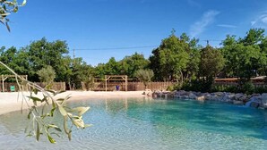 A clear pool with a sandy beach and a wooden structure in the background.