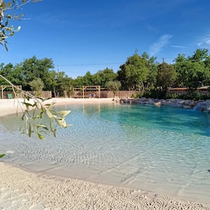 A clear pool with a sandy beach and a wooden structure in the background.