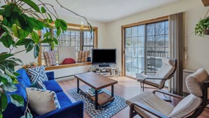 A living room with a blue sofa, a wooden coffee table, a TV, and a large window with a view of trees. - Game Room Condo, Sugar Hill, Pet-Friendly (Quechee)