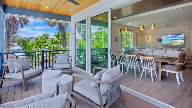 A spacious outdoor dining area with white furniture, a wooden ceiling, and large glass doors leading to a lush garden.
