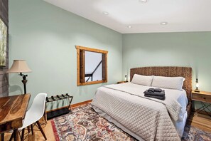 Un dormitorio con cama, escritorio, silla, espejo y alfombra estampada.