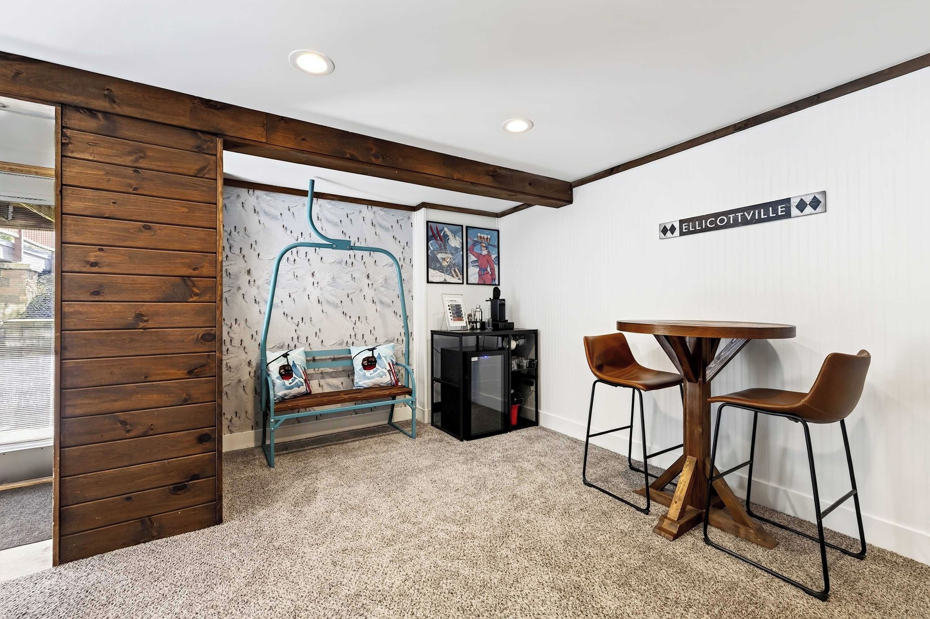 A room with a wooden bench, a small table with two chairs, a wall-mounted sign, and a ski-themed wallpaper.