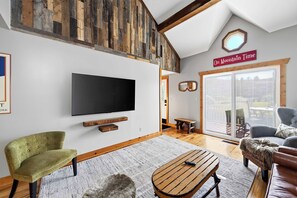 A modern living room with a large flat-screen TV, wooden beams, a bench, and a sign reading 'On Mountain Time'.