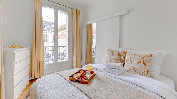 Una habitación de hotel con una cama, una bandeja con cruasanes y jugo de naranja, y vista a edificios por la ventana.