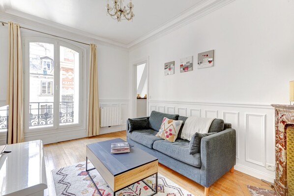 Un salon avec un canapé gris, une table basse noire et un tapis à motifs. On y trouve une grande fenêtre avec des rideaux, un lustre et un mur blanc orné de tableaux.
