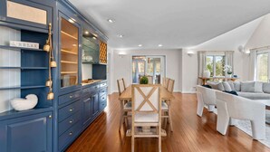 A dining area with a wooden table and chairs, a blue cabinet with glass doors, and a sofa in the background. - The Gathering Place - For 6 with Fire Pit (Mashpee)