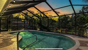 An indoor swimming pool with a glass ceiling, surrounded by tropical plants and a sunset view.