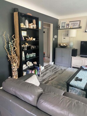 Una sala de estar con un sofá de cuero gris, una estantería llena de objetos diversos, un mueble gris y un televisor.