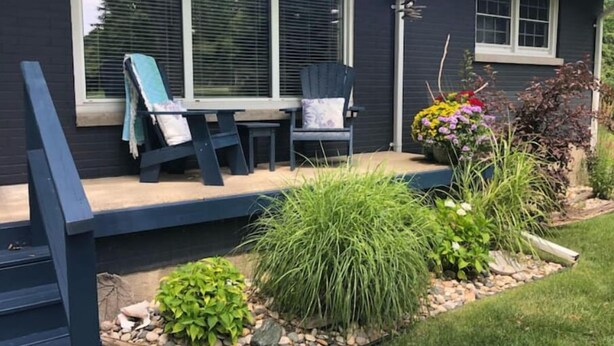 Une maison à l’extérieur sombre, avec un porche abrité comprenant une table et des chaises, et un jardin où poussent diverses plantes et fleurs.