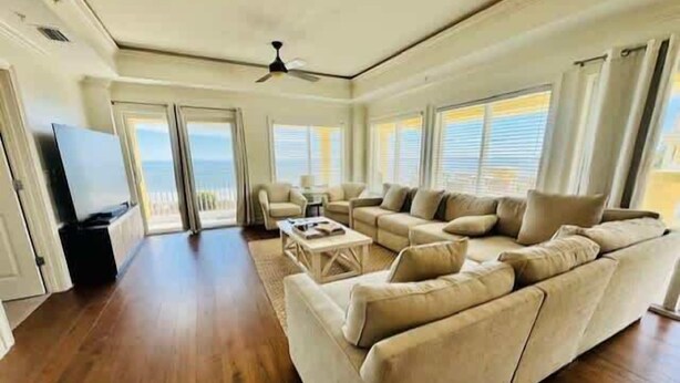 A spacious living room with a large sofa, a coffee table, and a TV. There are large windows with blinds, a ceiling fan, and a view of the ocean.