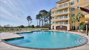 A swimming pool with a surrounding deck, a multi-story building with balconies, and a clear sky.