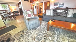 Ein gemütliches Wohnzimmer mit Sofa, Sessel und Couchtisch. Es gibt einen Essbereich mit Stühlen und einem Tisch. Der Raum verfügt über Holzbalken an der Decke und einen Teppich auf dem Boden.