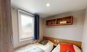 Un dormitorio pequeño y ordenado con una cama individual, una mesita de noche, una ventana con cortinas y un estante de madera sobre la cama con un baúl.