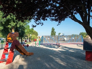 Un parque infantil con tobogán y columpios, rodeado de árboles y una cerca azul.