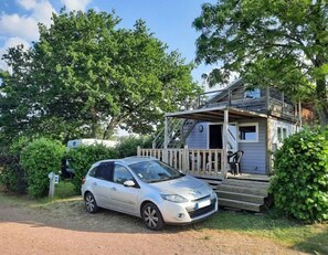 Une voiture blanche est garée devant une cabane rustique dotée d’une terrasse en bois et d’un porche abrité.