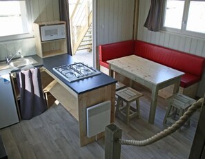 A compact kitchen with a gas stove, a sink, and a small refrigerator. There is a wooden table with a bench and a rope barrier. - ZEcamping 3* animé piscine chauffée locatif TV BBQ clim 36m2 6 pers (Argentonnay)
